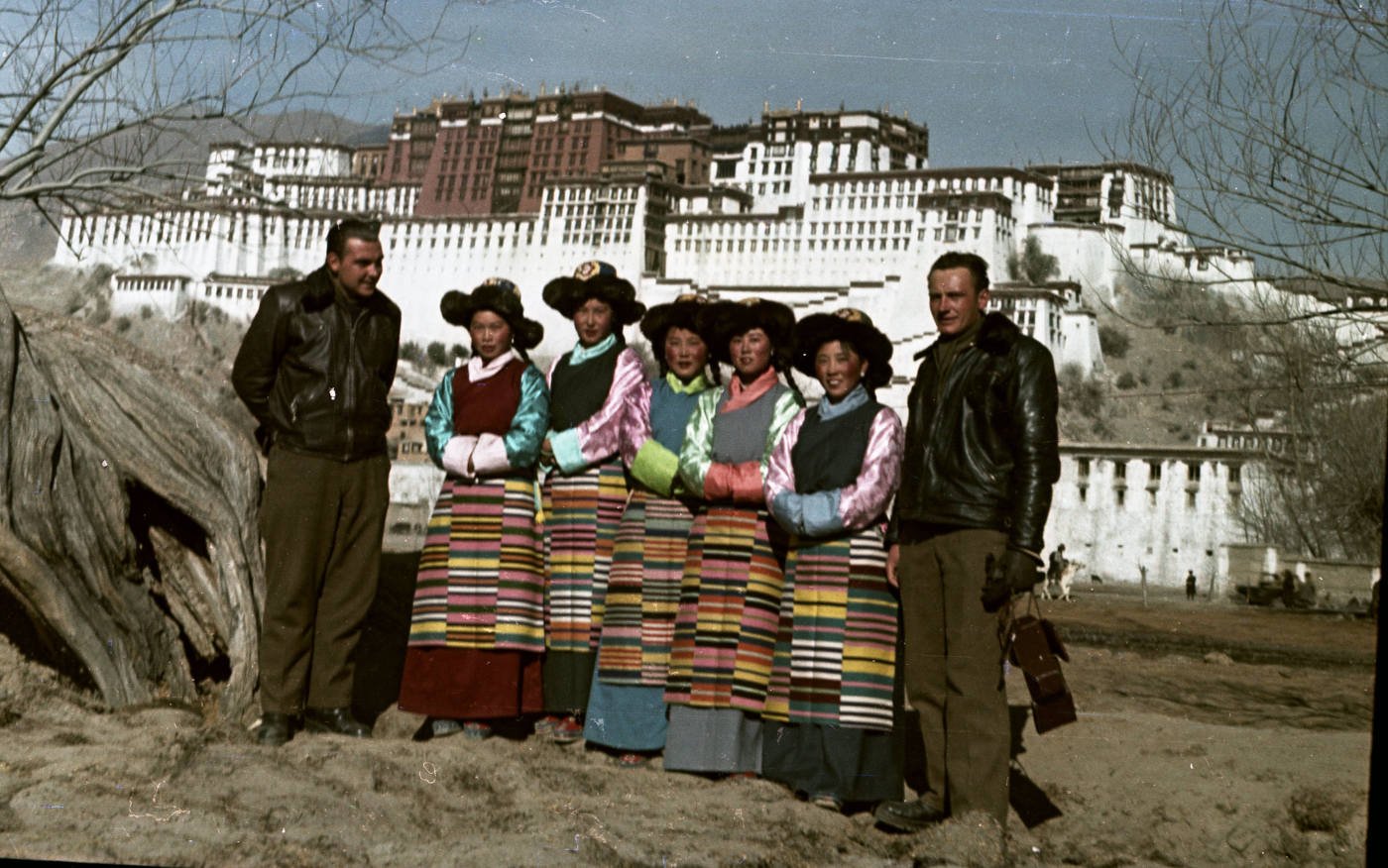 Skupinová fotografie s tibetskými ženami v národních krojích, Vladimír Sís vlevo, Josef Vaniš napravo. Fotografováno ve Lhase, v pozadí dalajlamovský palác Potala, 21. prosince 1954, publikováno se souhlasem držitele autorských práv Jana Vaniše.